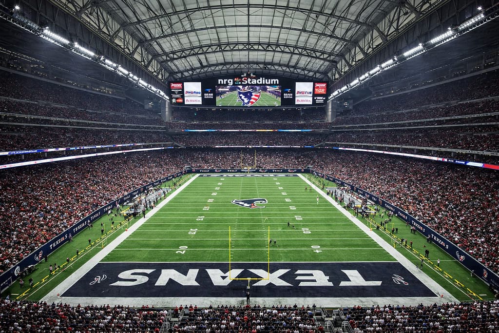 Wide-angle view of NRG Stadium in Houston, Texas, showing the Houston Texans football field under the closed retractable roof, with a packed crowd, bright green turf, end zone branding, and the stadiumβs massive video scoreboard during an NFL game.
